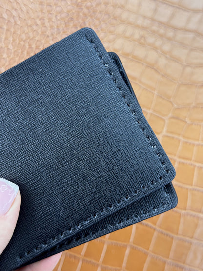 A close-up of a black textured cardholder wallet being held by a hand, showing fine stitching and a tan crocodile-embossed background.