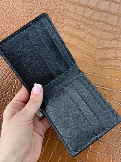 A black textured leather bifold wallet held open by a hand with light pink manicured nails, set against a brown crocodile-embossed leather background.