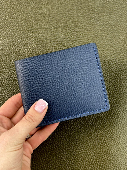 A navy blue textured leather bifold wallet held by a hand with light pink manicured nails, set against a dark olive green pebbled leather background.
