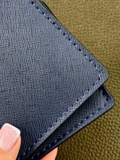 A close-up of a dark blue textured leather wallet or card holder with matching stitching, held by a hand with a light pink manicured fingernail, set against a textured olive green background.