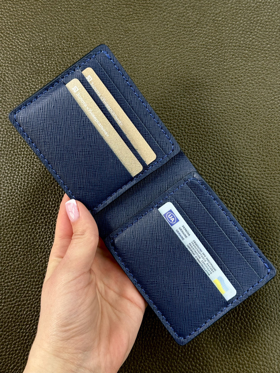 A navy blue textured leather bifold wallet held open by a hand, showing its interior card slots and ID pocket against a dark olive green textured background.