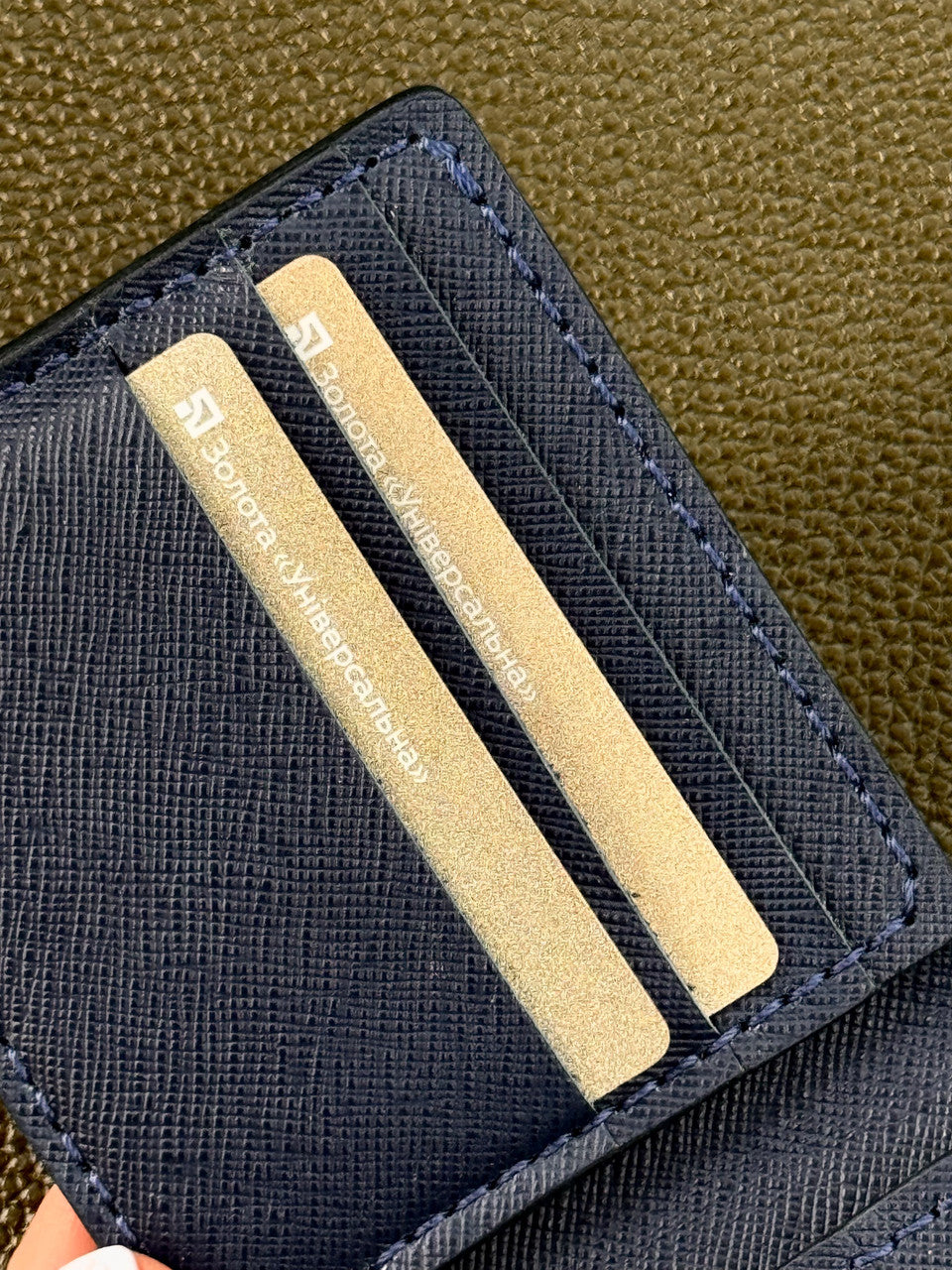 A close-up of a dark navy blue textured wallet or card holder with three gold-toned credit cards inserted, resting on an olive green textured surface.