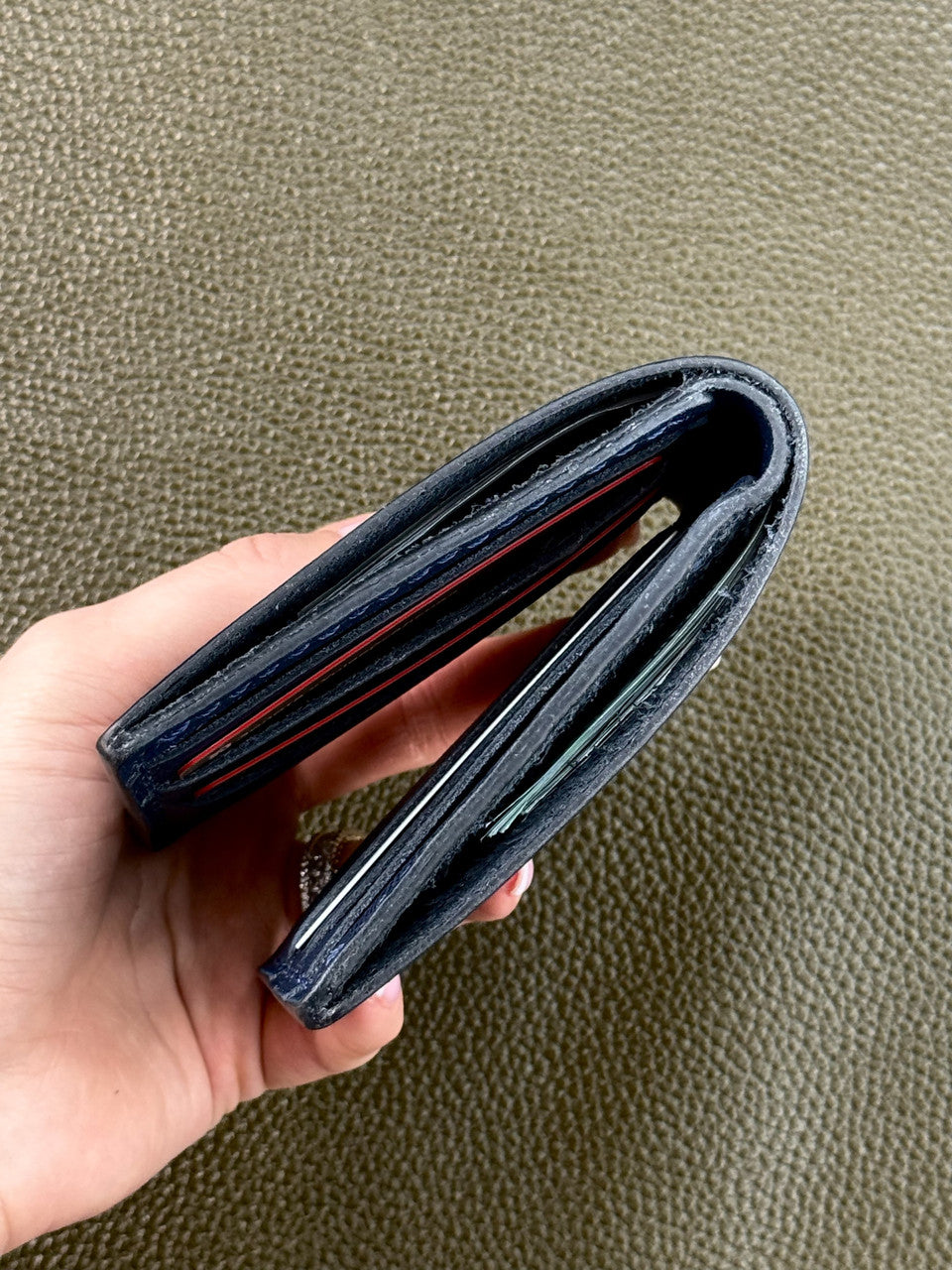A hand holding a dark navy blue leather wallet, showing its multiple card slots and interior lining, against a textured olive green background.