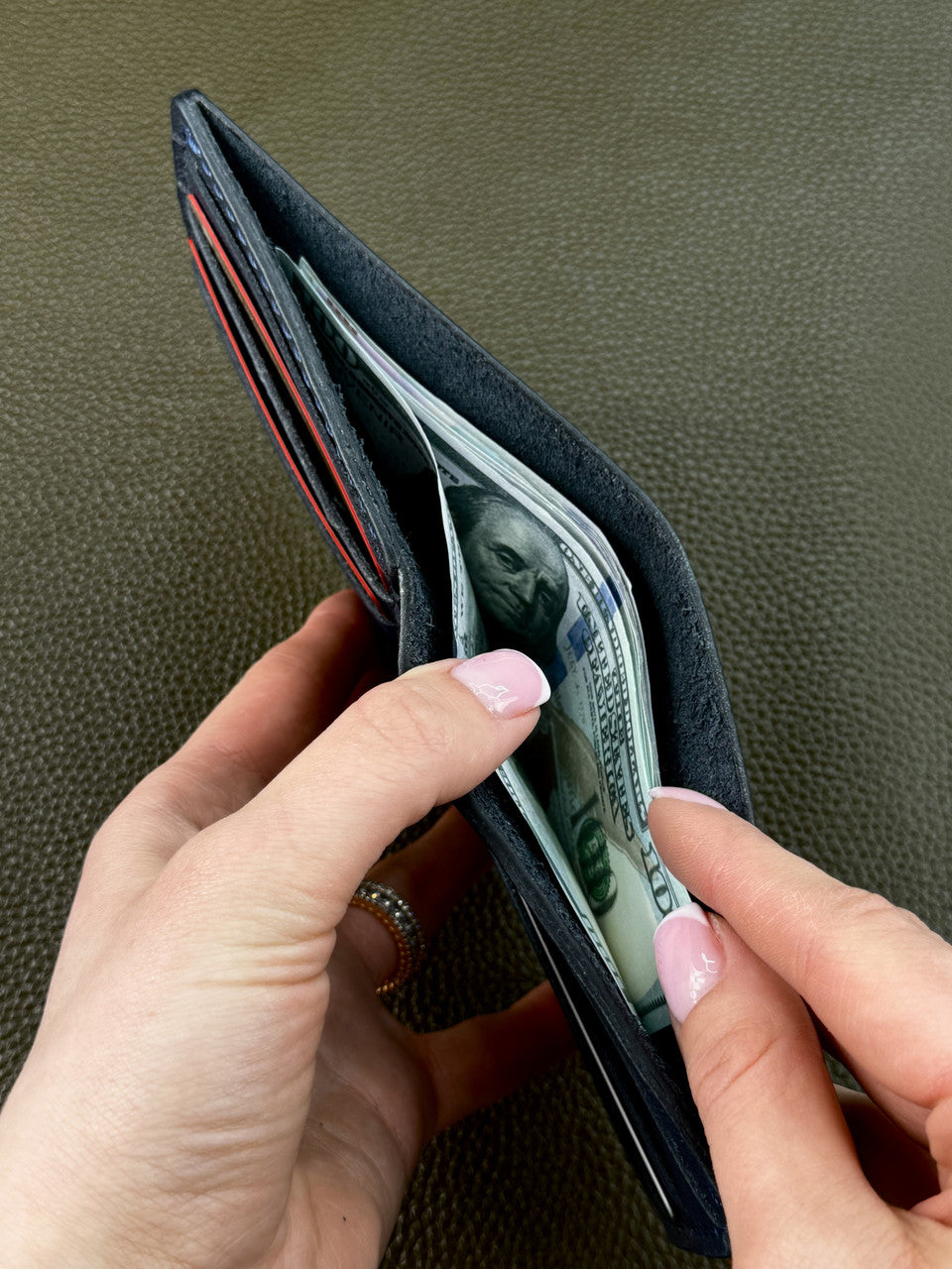 A close-up of a person's hands holding an open black leather wallet with cards and a dollar bill visible inside, set against a textured olive green background.