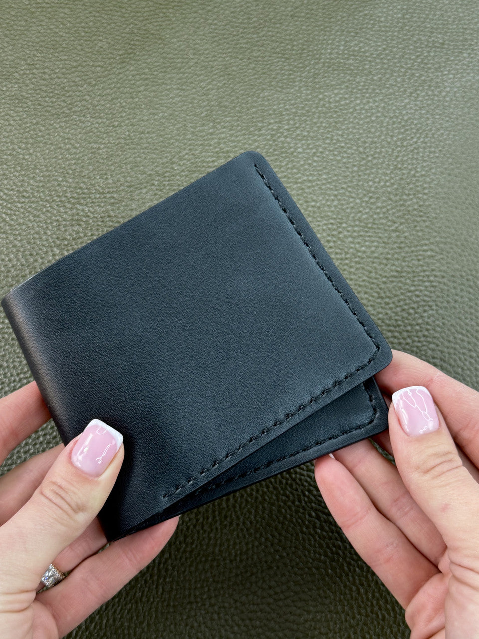 A black leather bifold wallet held by two hands against a textured olive green background.