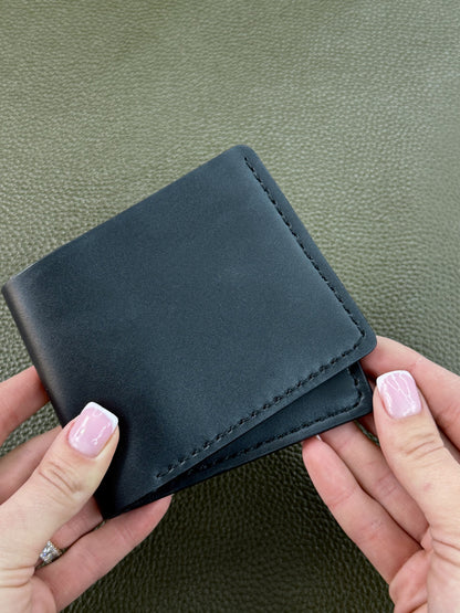 A black leather bifold wallet held by two hands against a textured olive green background.