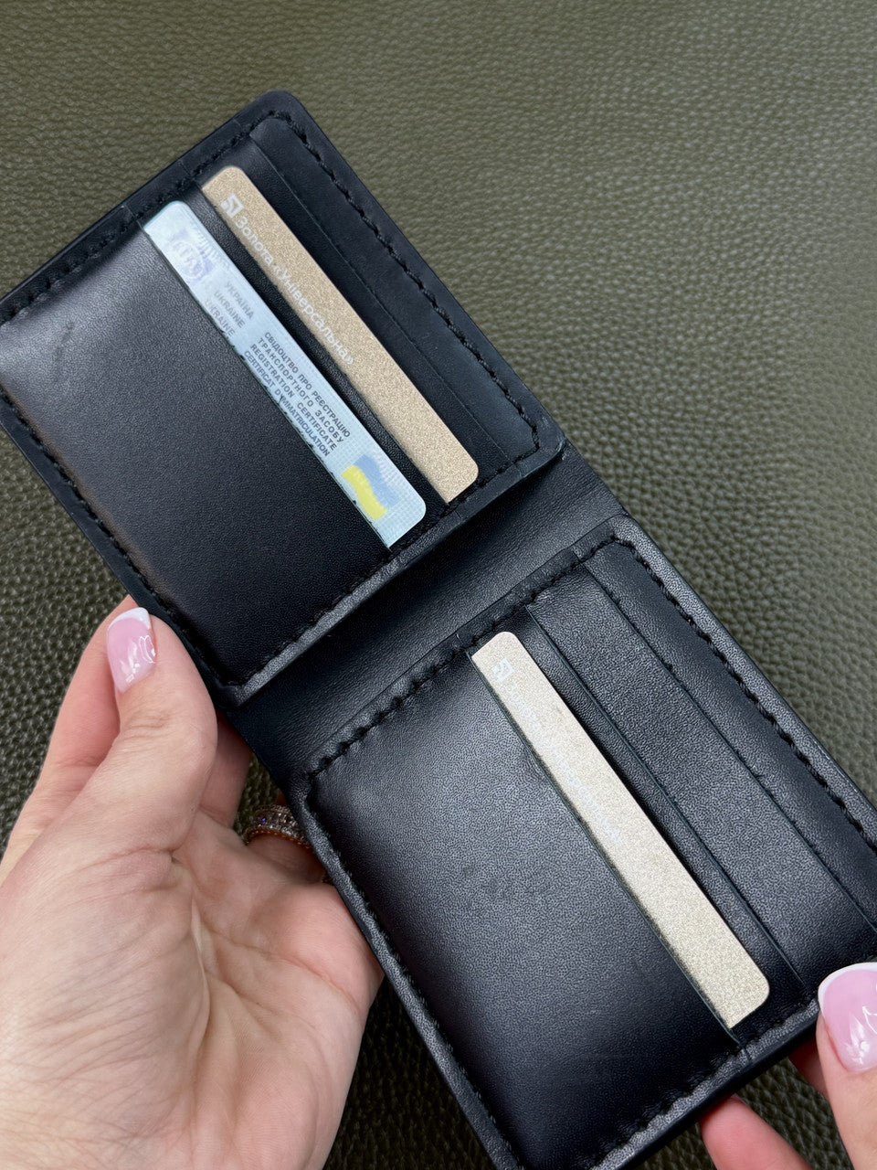 A black leather bi-fold wallet shown open, held by a hand with pink manicured nails, against a textured olive green background. The wallet contains several cards.