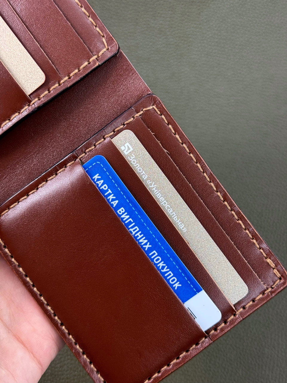 A close-up of an open brown leather bi-fold wallet held in a hand, showing card slots, contrast stitching, and sample credit cards inside.
