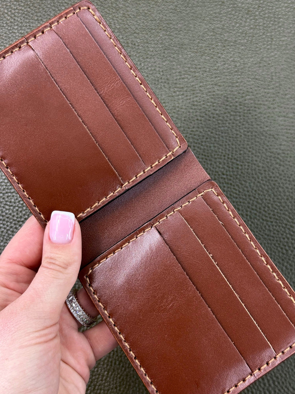 A brown leather bifold wallet held open by a hand with pink nail polish, showing multiple card slots and contrast stitching against a textured olive green background.