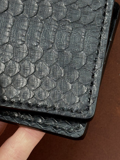 Close-up of a black textured leather item, likely a wallet or card holder, showing a scale-like embossed pattern and stitched edges, with a finger holding it against a brown background.