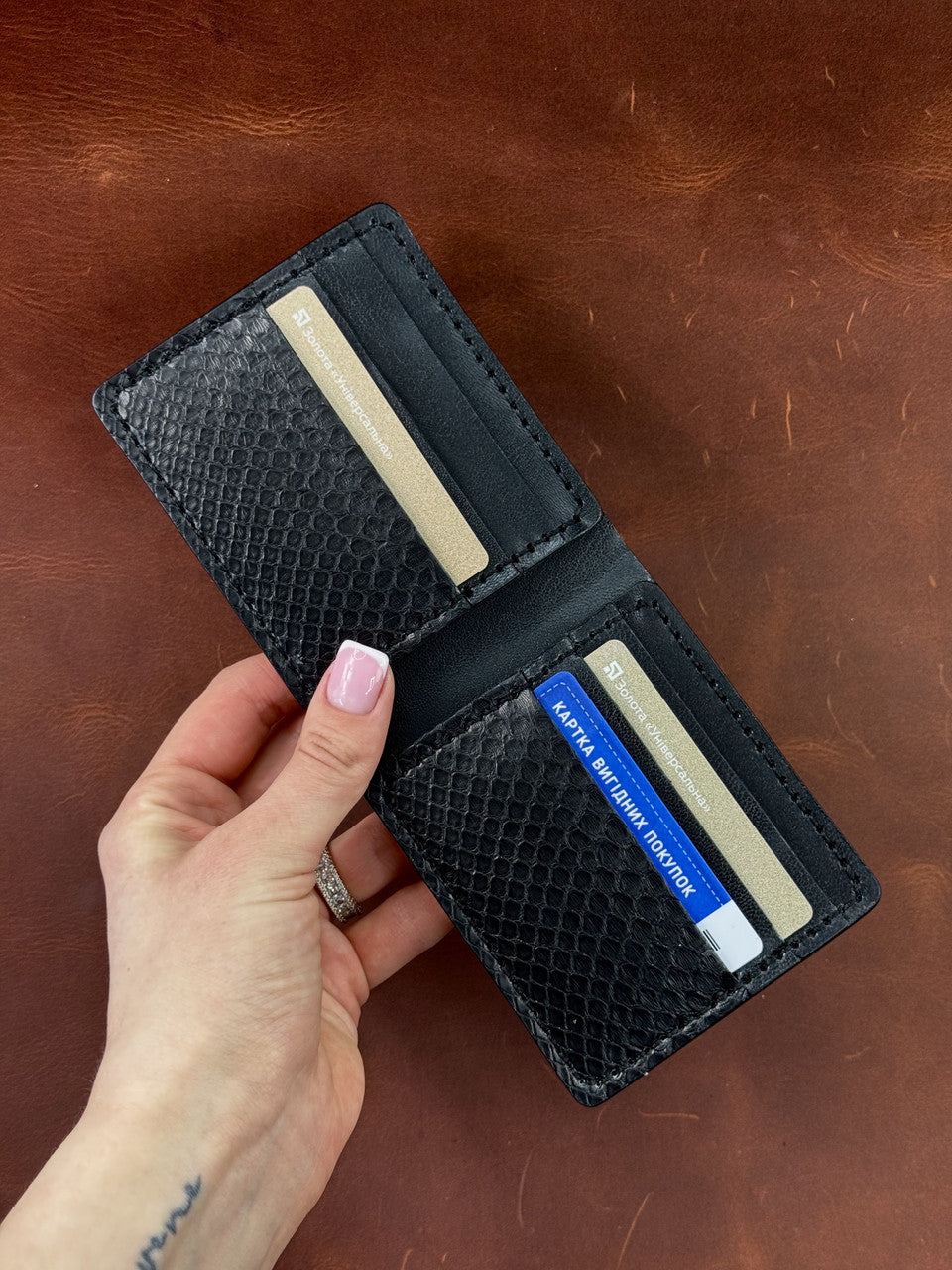 A black textured leather bifold wallet held open by a hand, showing card slots with sample cards inside, set against a brown leather background.
