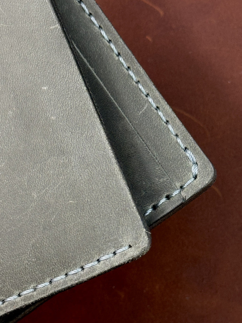 Close-up of a gray leather wallet corner showing white contrast stitching on a dark brown surface.