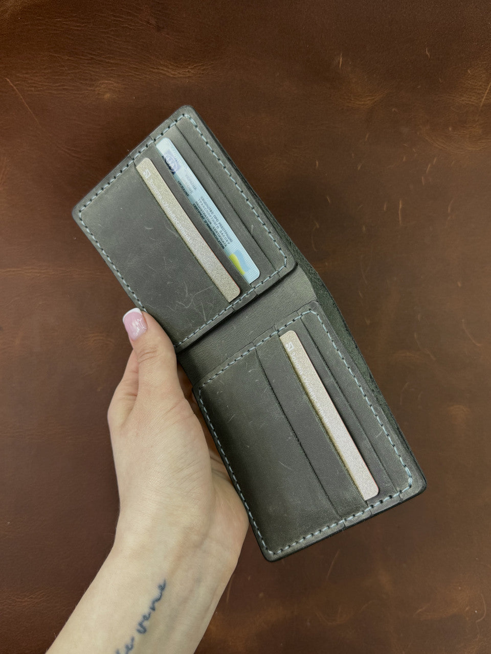 A dark gray leather bi-fold wallet held open by a hand, showing multiple card slots with credit cards inserted, set against a brown leather background.