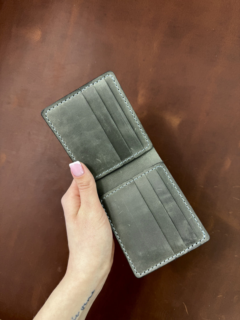 A person's hand with pink nail polish holds an open dark gray leather bifold wallet with multiple card slots, set against a brown leather background.