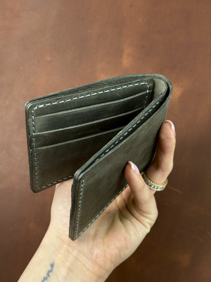 A dark olive green leather bifold wallet held open in a hand, showing its interior card slots and white contrast stitching against a brown leather background.