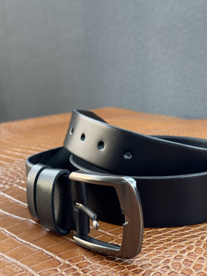 A black leather belt with a silver-tone metal buckle, coiled on a textured brown surface against a dark gray background.