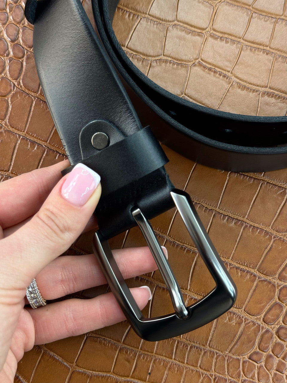 A close-up of a black leather belt with a silver-tone metal buckle, held by a hand with pink manicured nails over a brown crocodile-embossed textured surface.