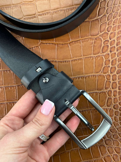 A close-up of a black leather belt with a silver-toned metal buckle, held by a hand with pink manicured nails against a brown crocodile-embossed textured background.