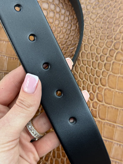 A close-up of a hand holding a black leather belt with silver-tone metal rivets and adjustment holes, set against a textured tan crocodile-patterned background.