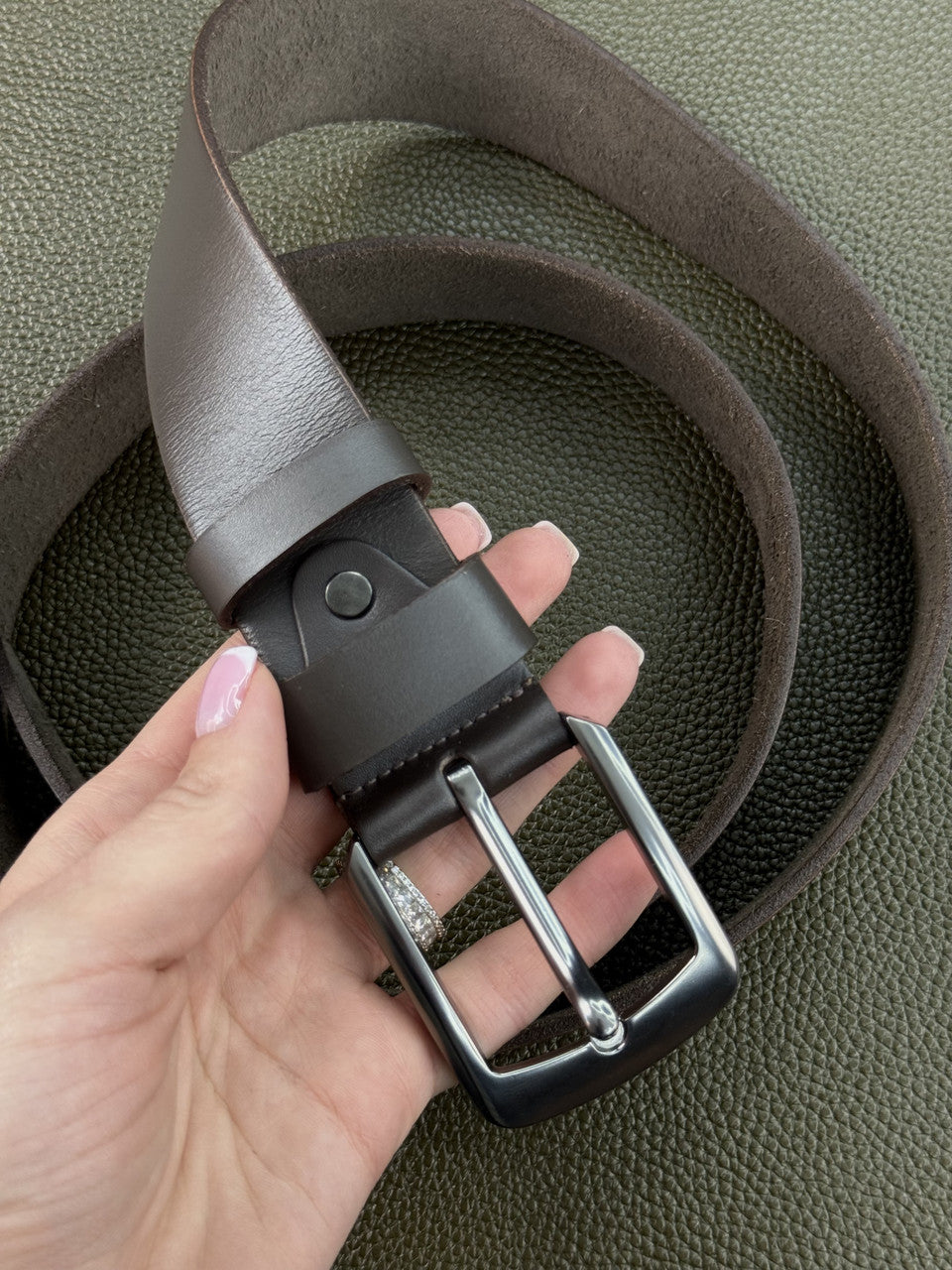 A dark brown leather belt with a black keeper and silver-tone buckle, held by a hand over a textured olive green background.