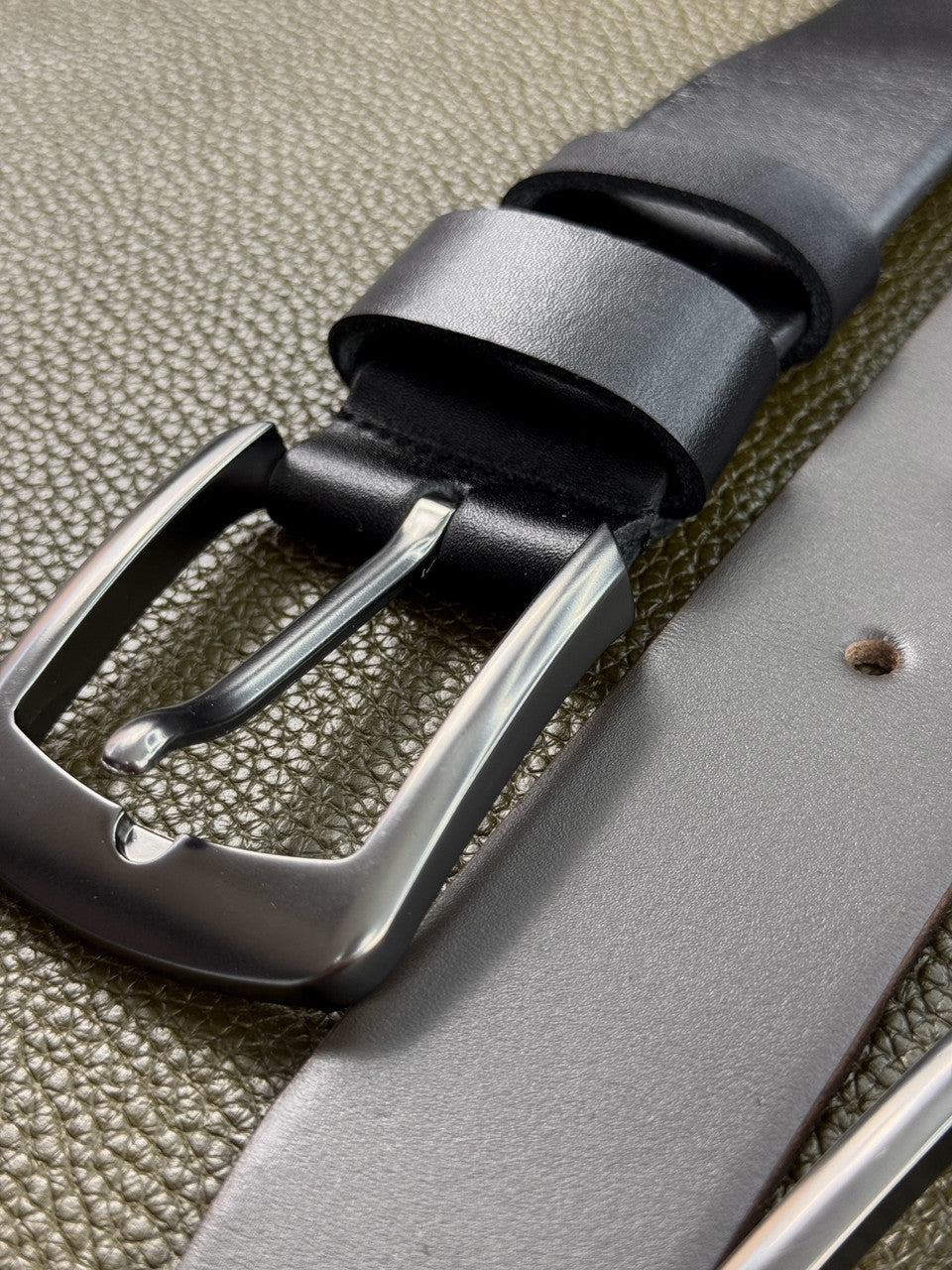 A close-up of a dark gray leather belt with a polished silver-tone metal buckle, shown resting on a textured olive green surface.