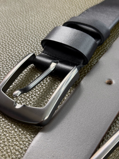 A close-up of a dark gray leather belt with a polished silver-tone metal buckle, shown resting on a textured olive green surface.