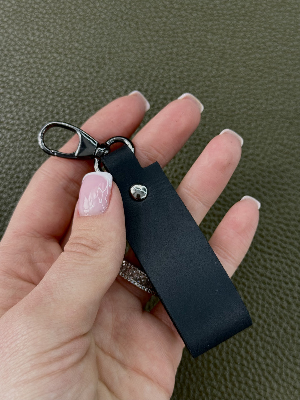 A close-up of a hand holding a black leather keychain strap with a silver-tone metal clasp and a small clear rhinestone accent, set against a textured olive green background.