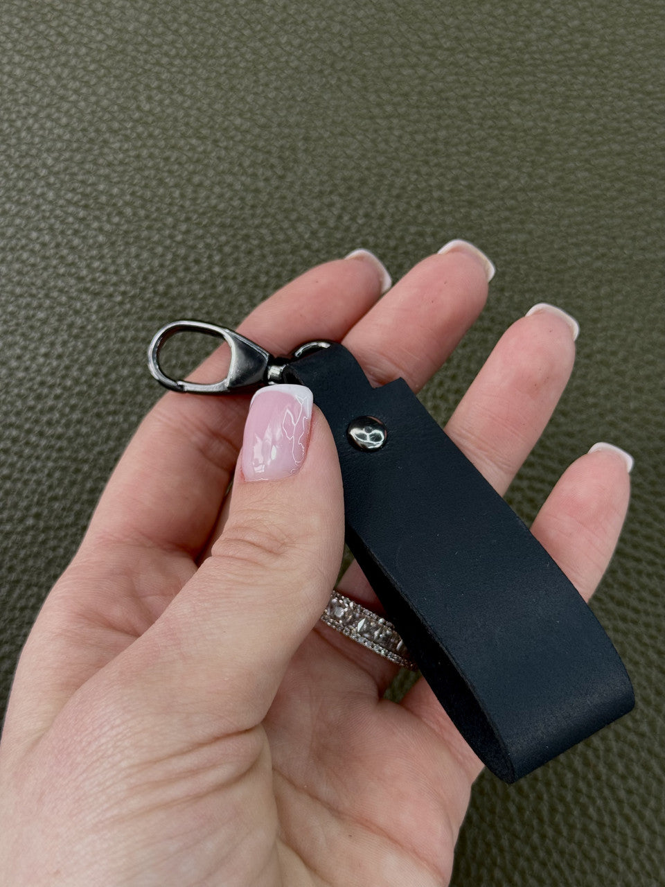 A close-up of a hand holding a black leather keychain with a silver metal lobster clasp and a small silver skull emblem, set against a textured olive green background.