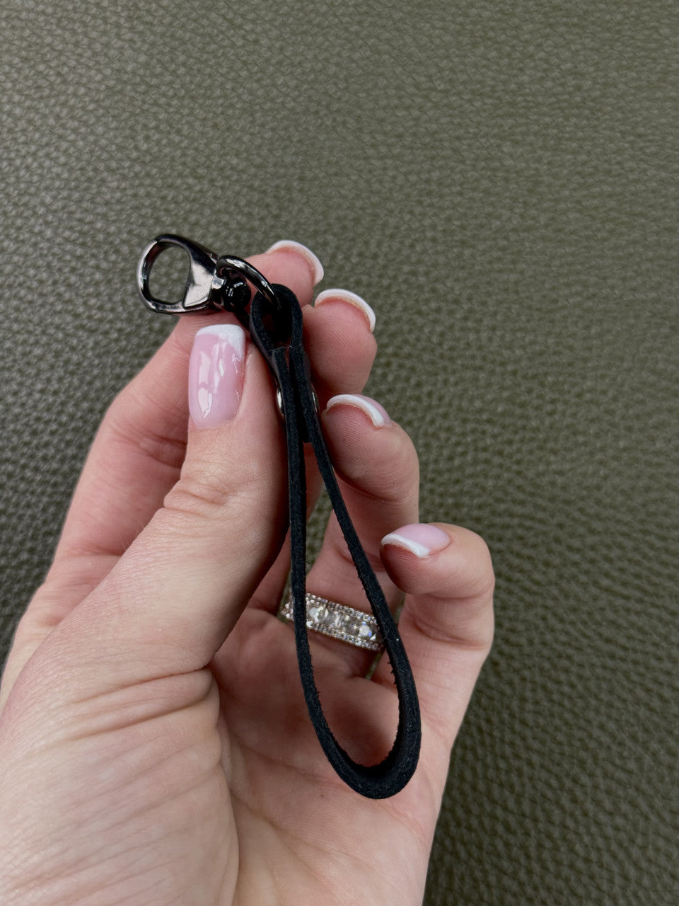 A close-up of a hand holding a black fabric wrist strap keychain with a silver-tone lobster clasp, set against a textured olive green background.