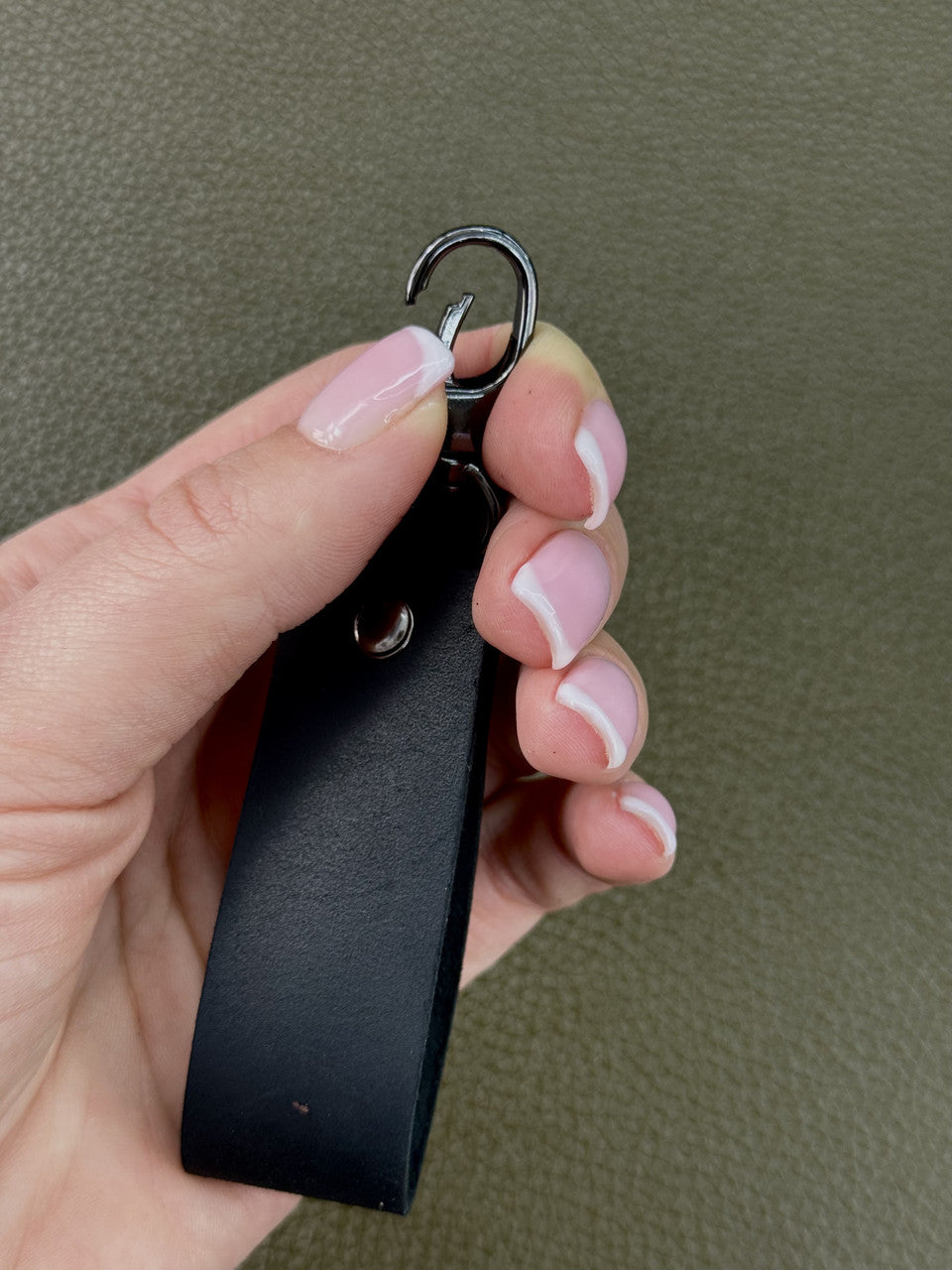 A close-up of a hand holding a black leather keychain with a silver metal split ring, set against a textured olive green background.