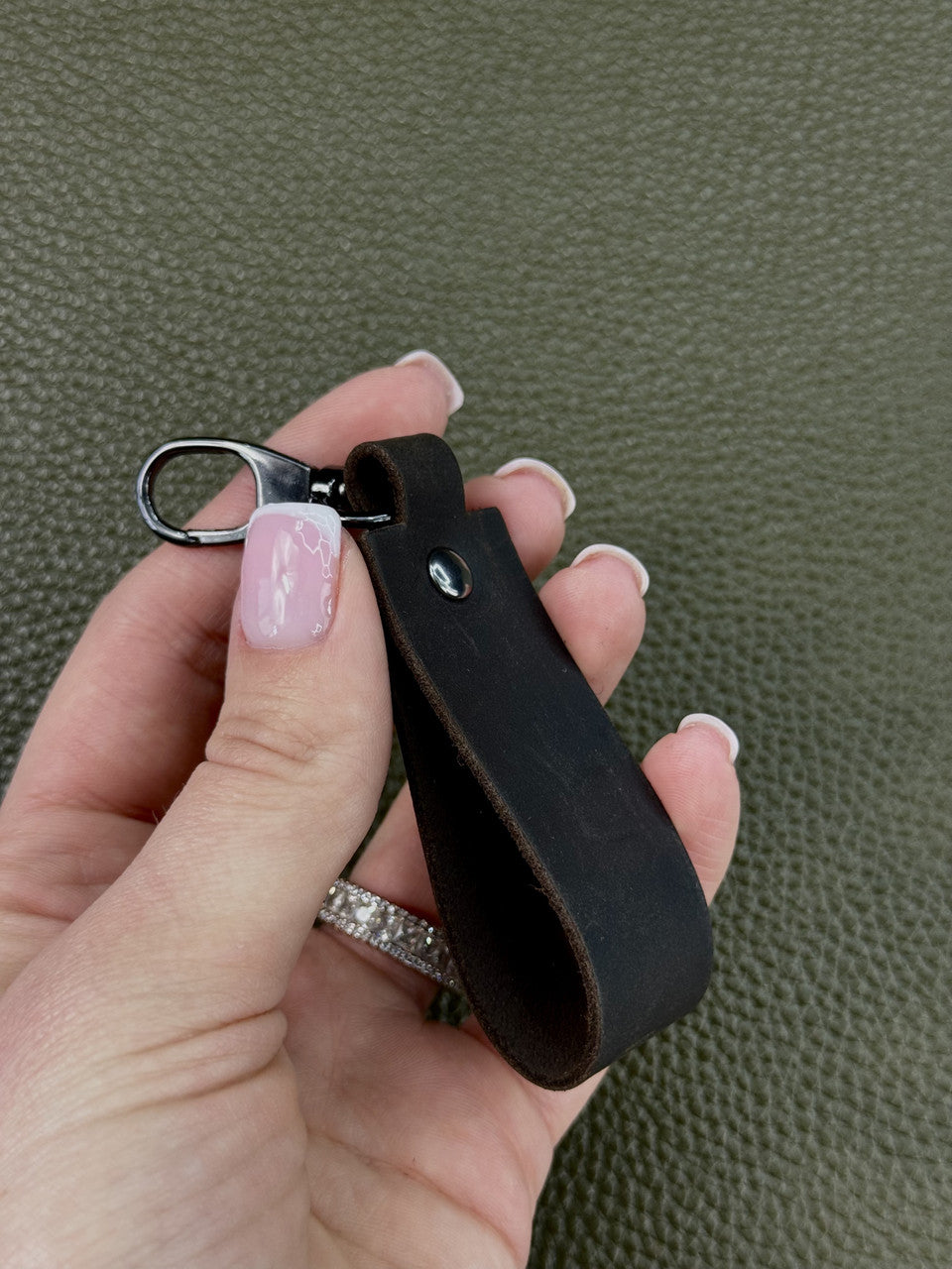 A close-up of a hand holding a black leather keychain strap with a silver metal lobster clasp, set against a textured olive green background.