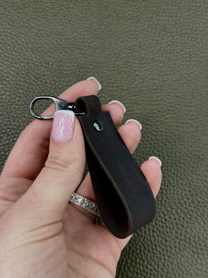 A close-up of a hand holding a black leather keychain strap with a silver metal lobster clasp, set against a textured olive green background.
