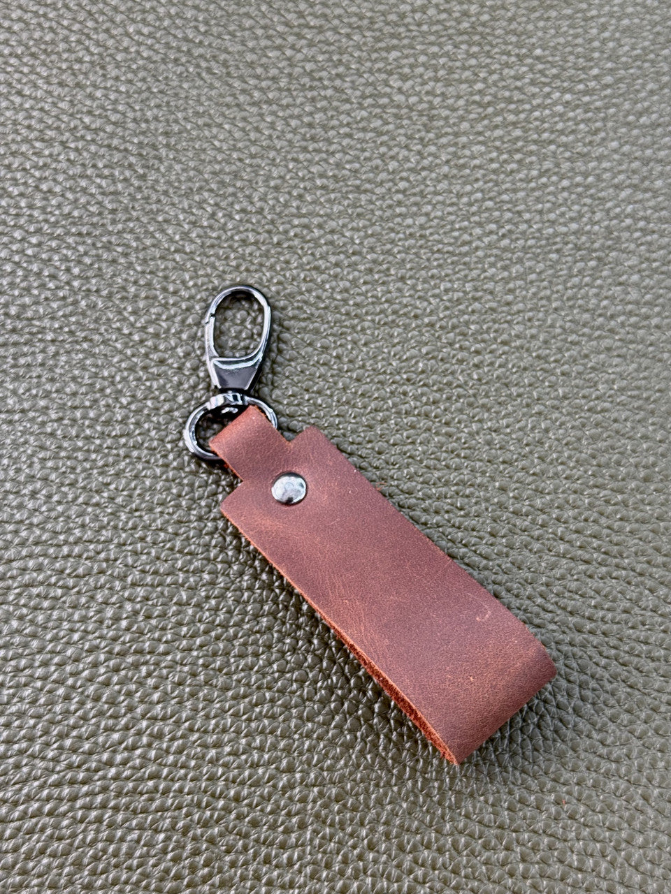 A brown leather keychain tag with a silver metal lobster clasp, shown on a textured olive green surface.