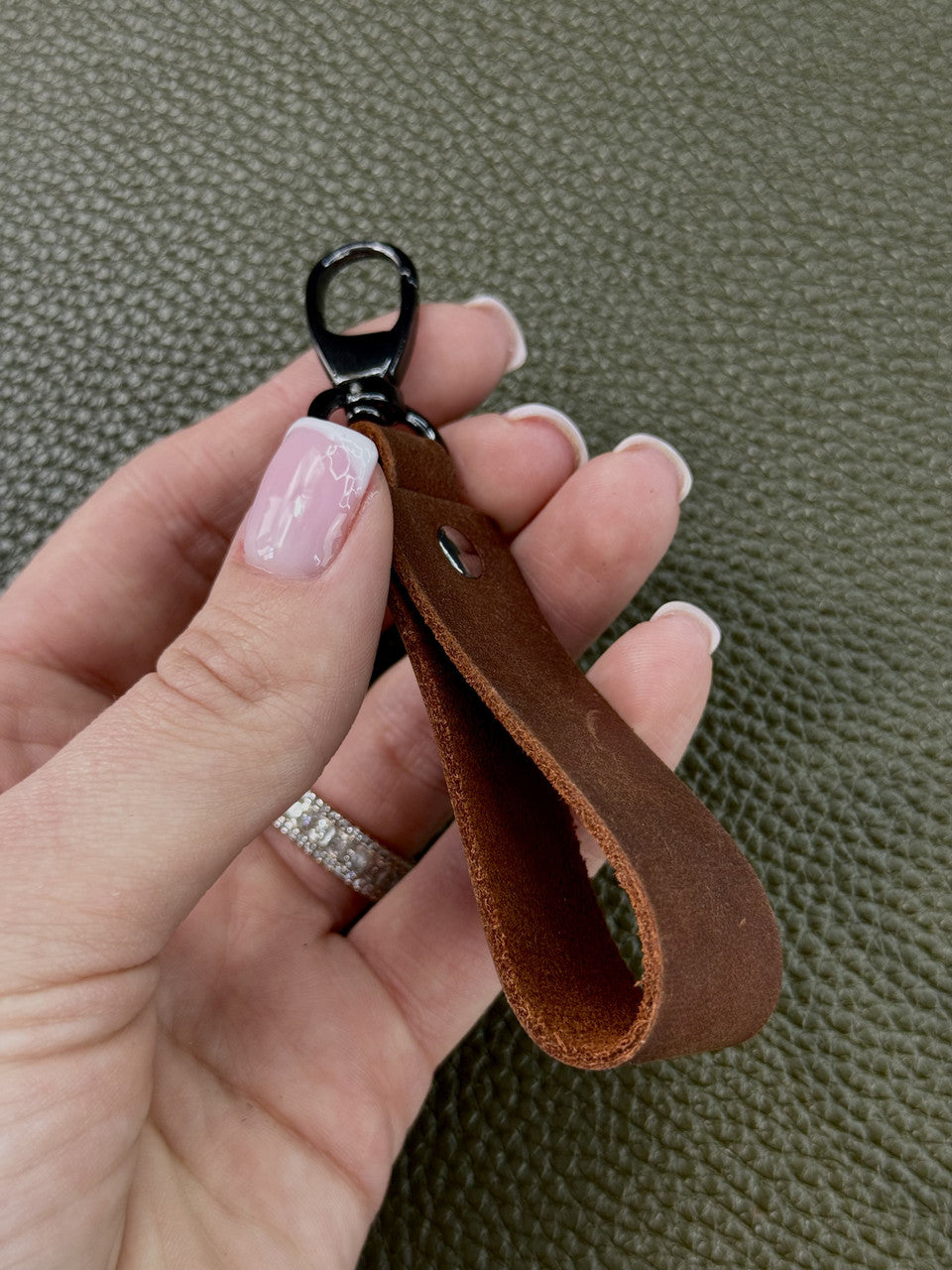 A close-up of a hand holding a brown leather keychain loop with a black metal clasp, set against a textured olive green background.