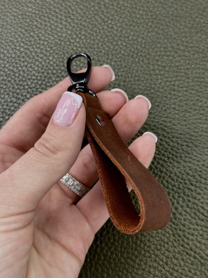 A close-up of a hand holding a brown leather keychain loop with a black metal clasp, set against a textured olive green background.