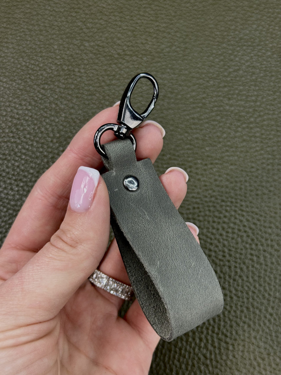 A hand with pink manicured nails holds a dark gray leather keychain with a black swivel clasp against a textured olive green background.