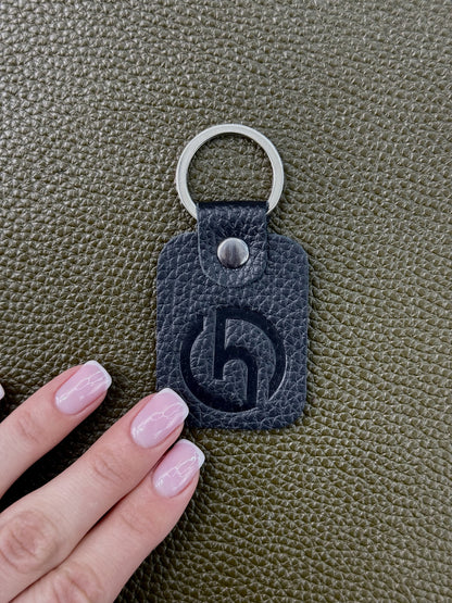 A dark gray pebbled leather key tag with a silver metal key ring and a debossed stylized 'h' logo, shown next to a hand with light pink manicured nails on a textured olive green surface.