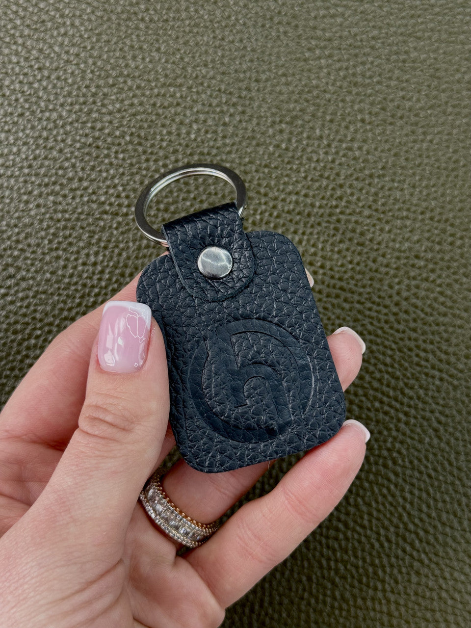 A hand holding a small black textured leather keychain with a silver metal key ring and snap button, set against a dark olive green textured background.