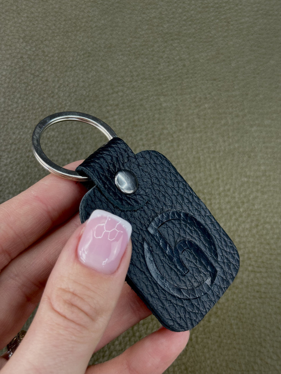 A close-up of a hand holding a black textured leather keychain with a silver metal key ring and a small silver snap button. The keychain features an embossed circular logo on the front, set against a textured olive green background.