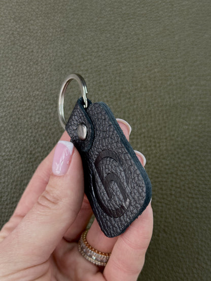 A hand with pink manicured nails holds a black textured leather keychain featuring an embossed stylized 'C' logo and a silver split ring, set against a textured olive green background.