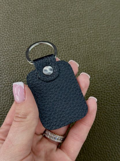 A close-up of a hand holding a small black textured leather keychain with a silver metal key ring and snap button, set against a dark olive green textured background.