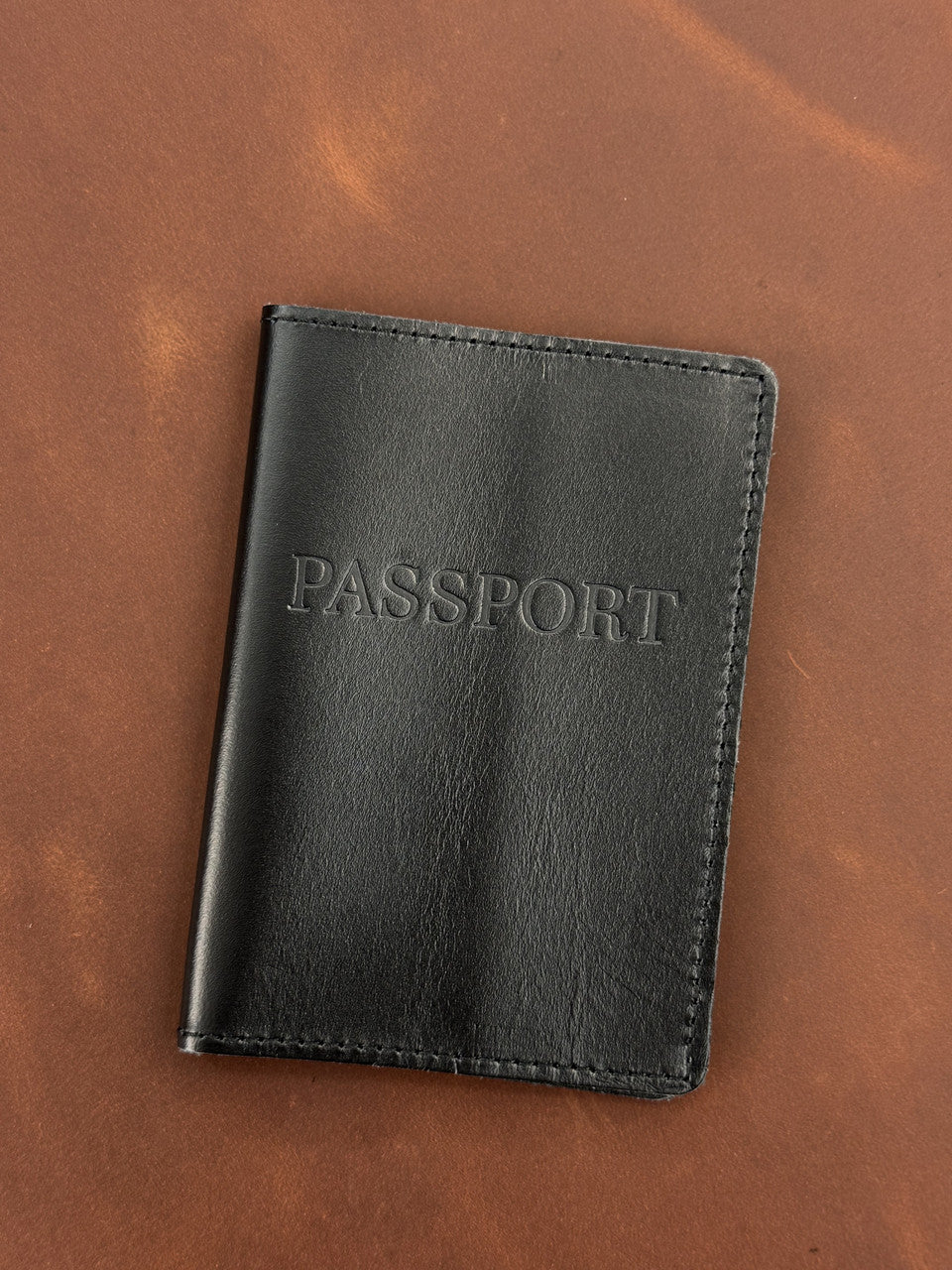 A black leather passport cover with 'PASSPORT' embossed on the front, displayed on a brown leather background.