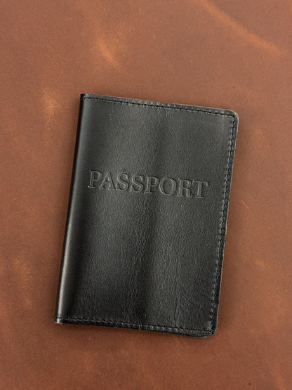 A black leather passport cover with 'PASSPORT' embossed on the front, displayed on a brown leather background.