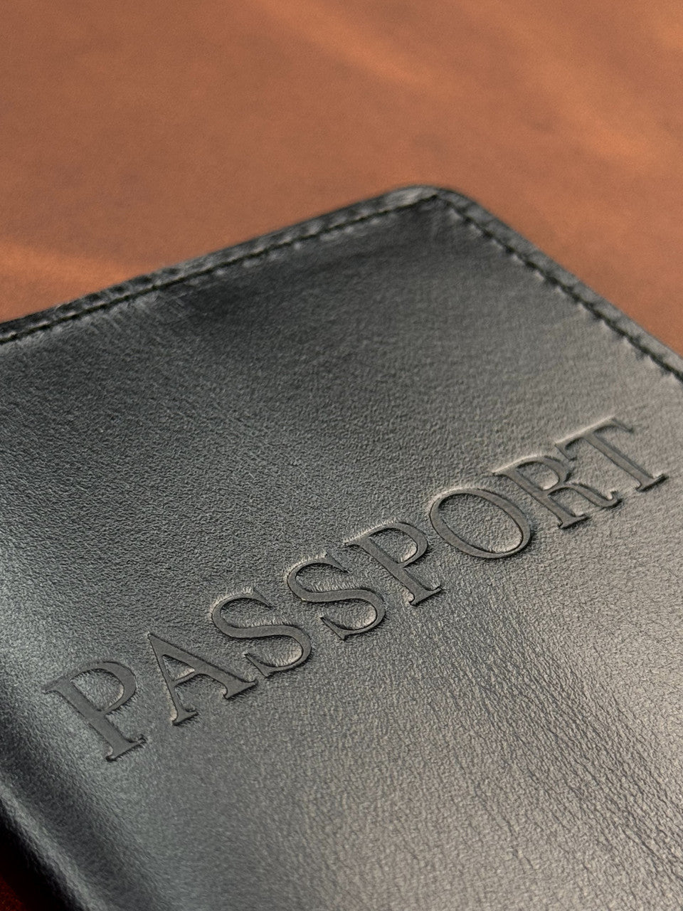 Close-up of a black leather passport cover with the word 'PASSPORT' embossed on the front, resting on a brown leather surface.