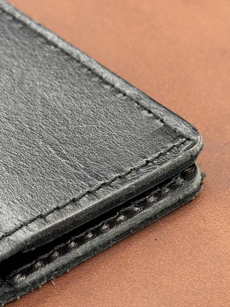 Close-up of a black leather wallet corner showing stitched edges and textured leather grain against a warm brown background.
