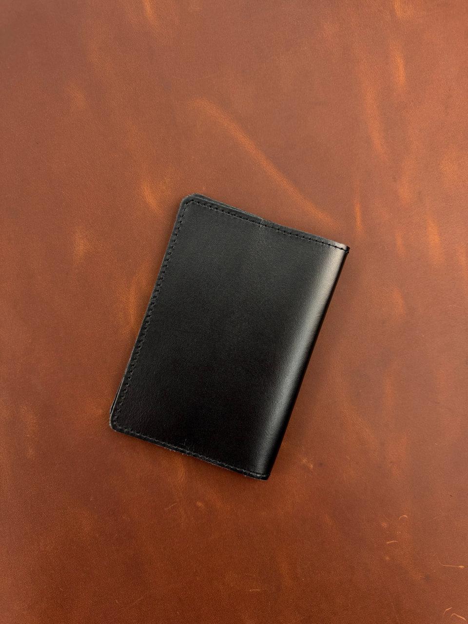 A black leather card holder wallet with visible stitching, placed diagonally on a textured brown leather background.
