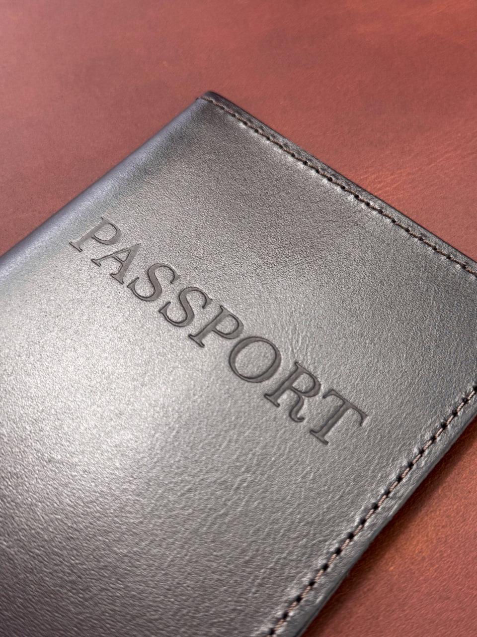 A close-up of a dark gray textured leather passport holder with the word 'PASSPORT' embossed on the front, resting on a brown wooden surface.