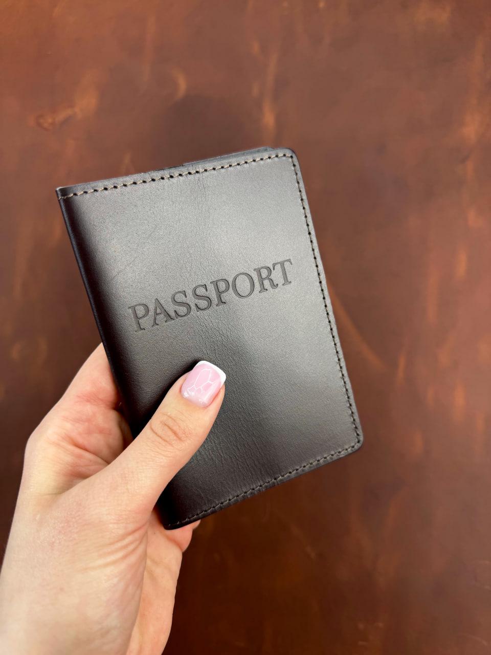 A hand holding a black leather passport holder with the word 'PASSPORT' embossed on the front, set against a brown textured background.