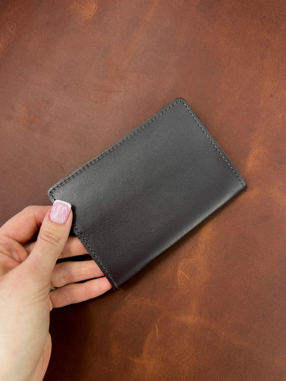 A hand with light pink nail polish holds a slim black leather card holder against a textured brown leather background.