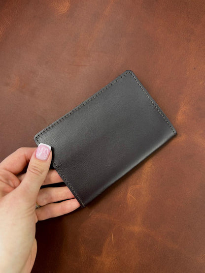 A hand with light pink nail polish holds a slim black leather card holder against a textured brown leather background.